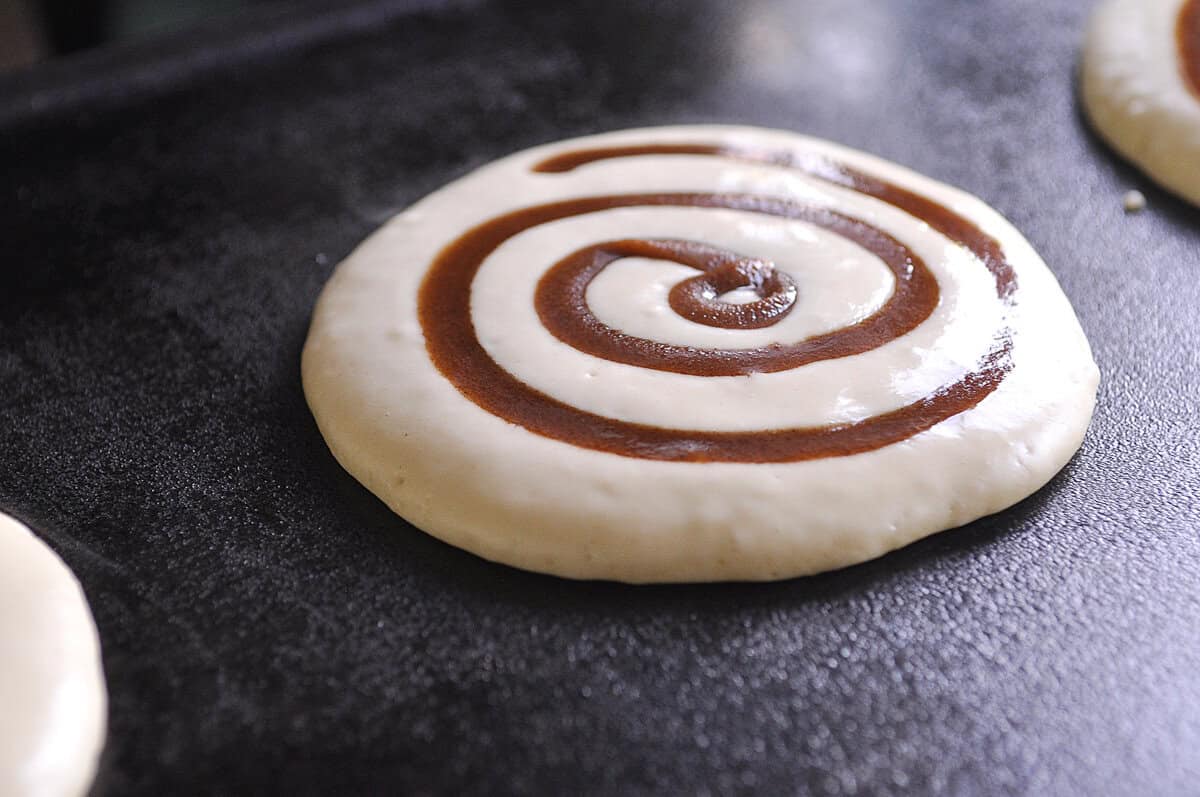 cinnamon roll pancake batter on griddle with cinnamon swirl in it